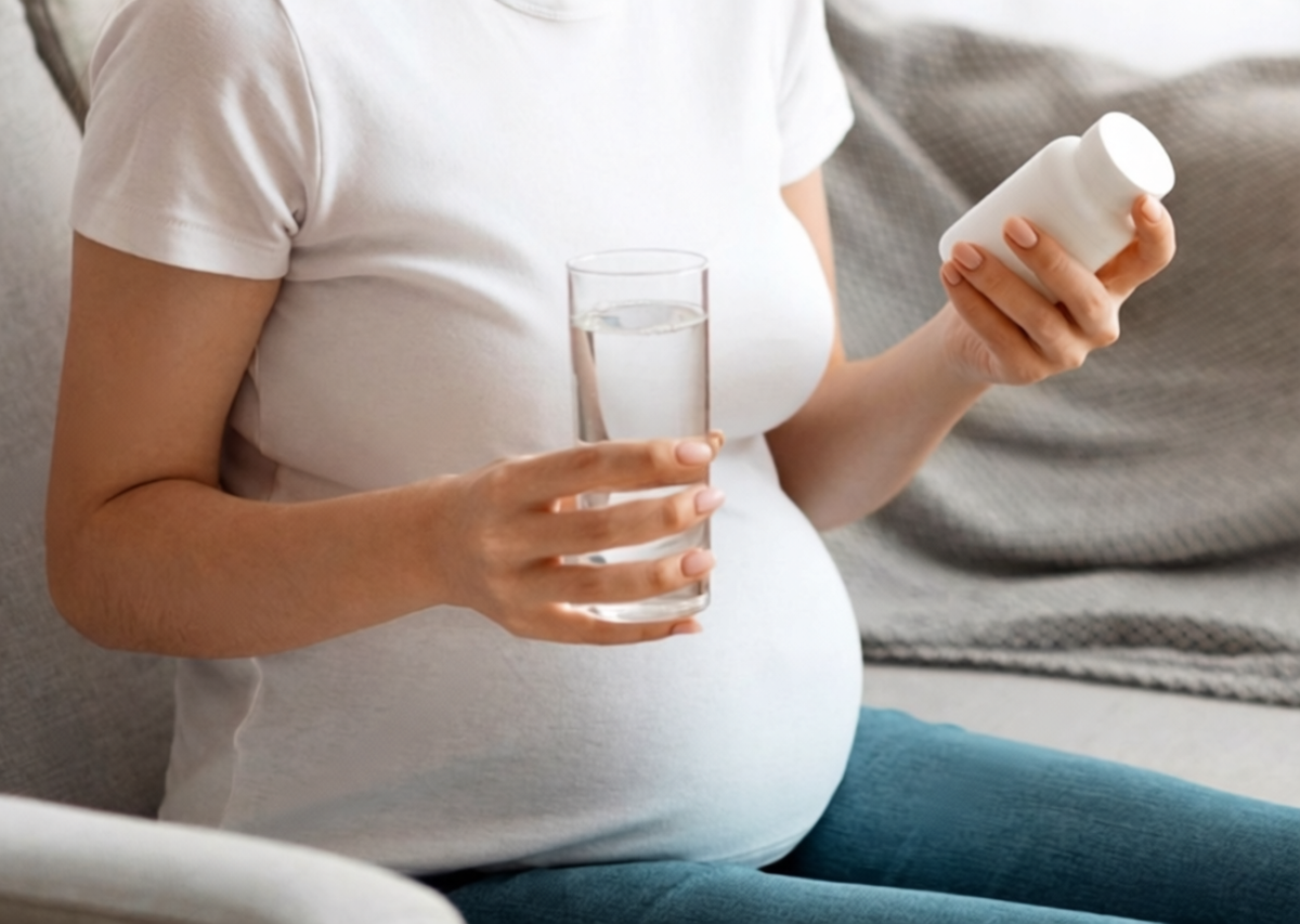 Close-up of a pregnant woman holding a bottle of supplements and a glass of water, reading the nutritional information on the label as she hesitates over whether to take the supplement before consuming it